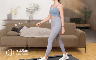 A woman walking on a treadmill in a living room, with a person sleeping on the couch behind her.